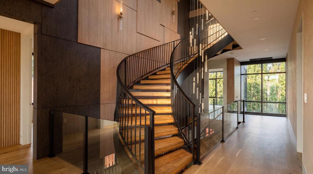 Curved wood staircase with black railings and glass balustrades in a modern, sunlit foyer.