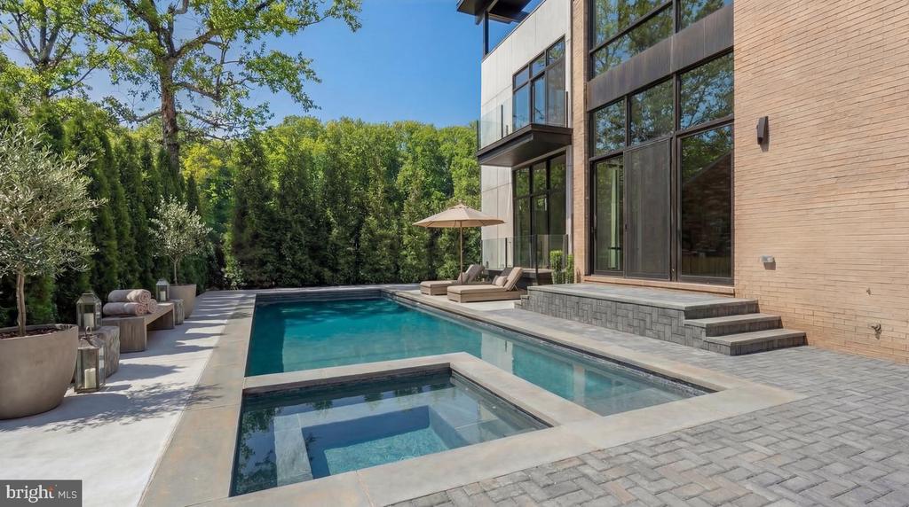 Outdoor pool and spa beside a modern brick-and-glass home, with a paved patio and lounge seating.