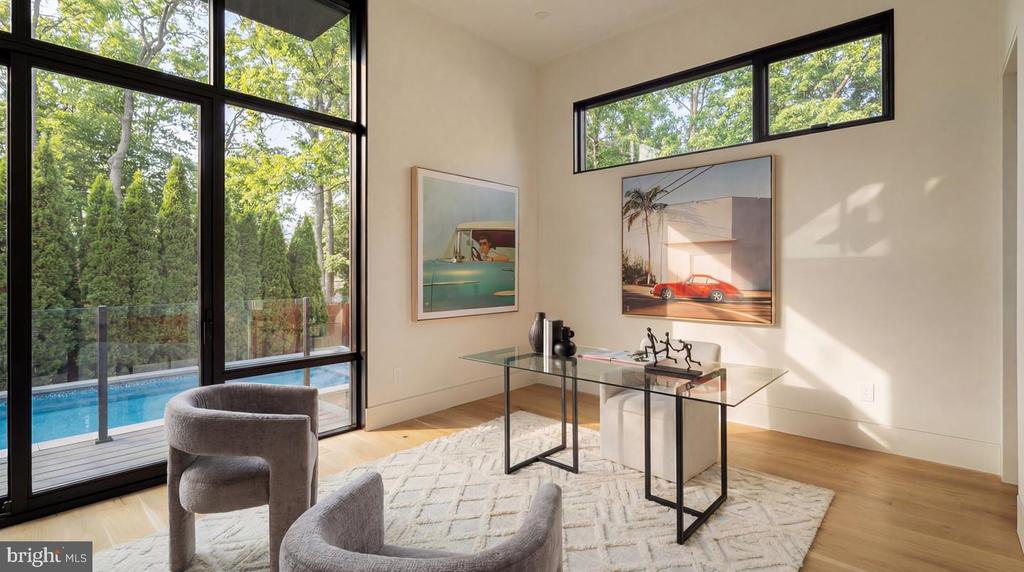 Furnished sitting room with tall windows overlooking a swimming pool and leafy trees.