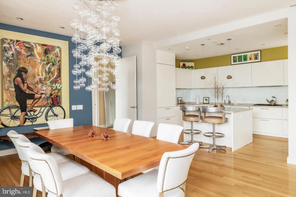 Dining room with a long wooden table and white chairs beside a bright, open kitchen.