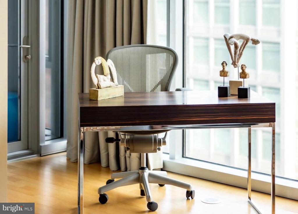 Home office with a dark wood desk, rolling chair, and sculptures beside tall windows.