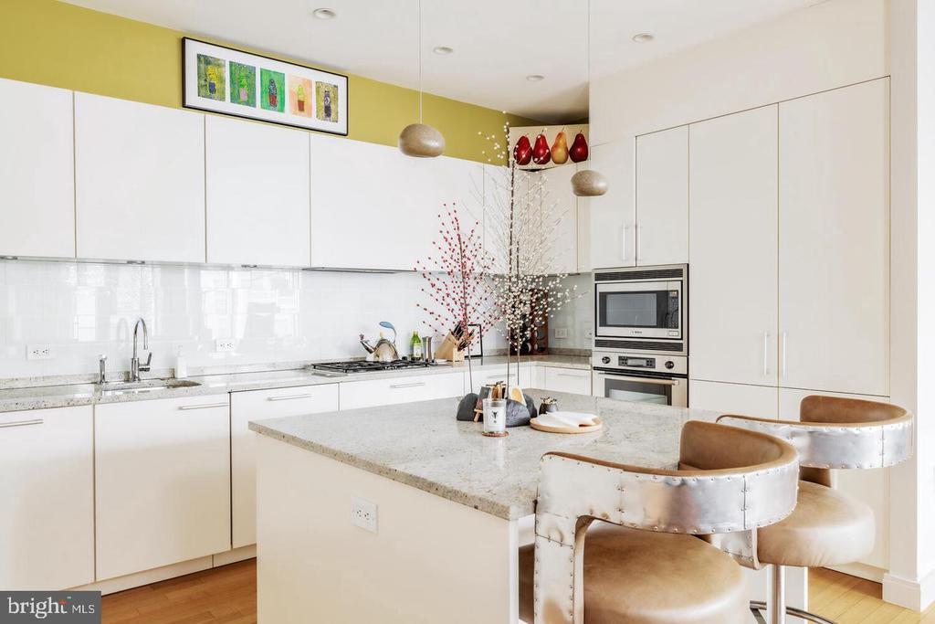Modern white kitchen with a stone island, upholstered stools, and a mustard-yellow accent wall.