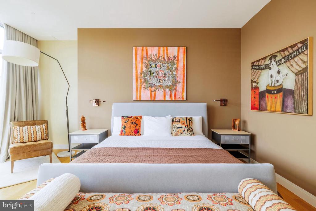 Bedroom with a tan accent wall, colorful artwork, and a chair beside the window.