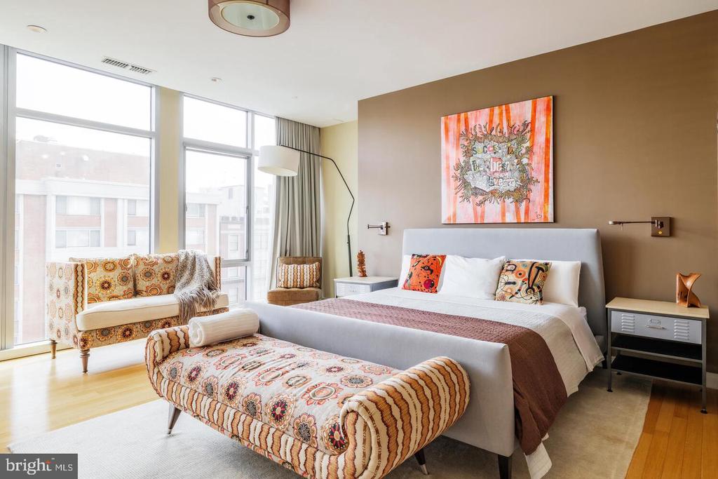 Furnished bedroom with a large bed, patterned bench and loveseat, and tall windows.