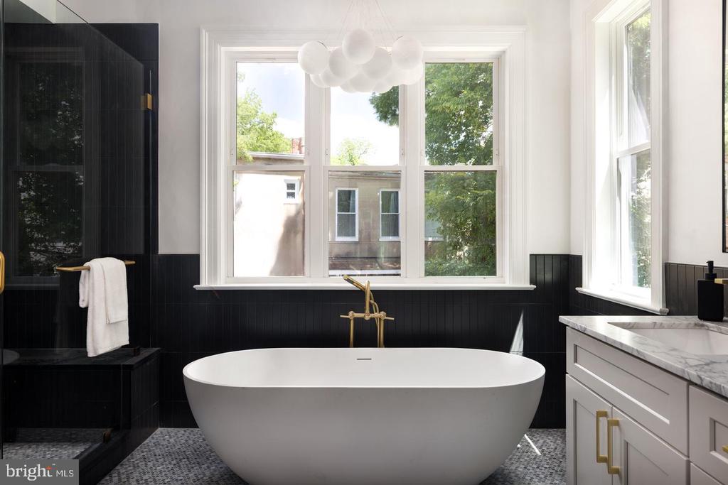 Bathroom with a freestanding soaking tub beneath large windows, black tile, and a marble-topped vanity.