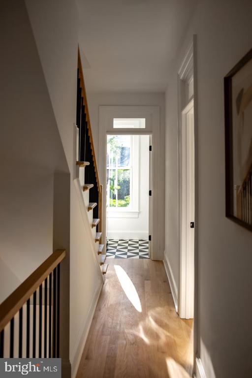 Hallway with wood floors, staircase, checkerboard tile, and sunlight through a rear window.