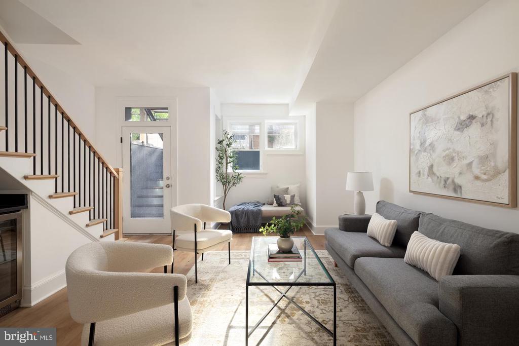 Staged living room with a gray sofa, cream chairs, glass coffee table, and staircase.