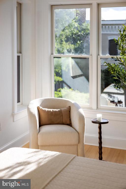 Staged bedroom with a cream bed and armchair beside tall sunlit windows.