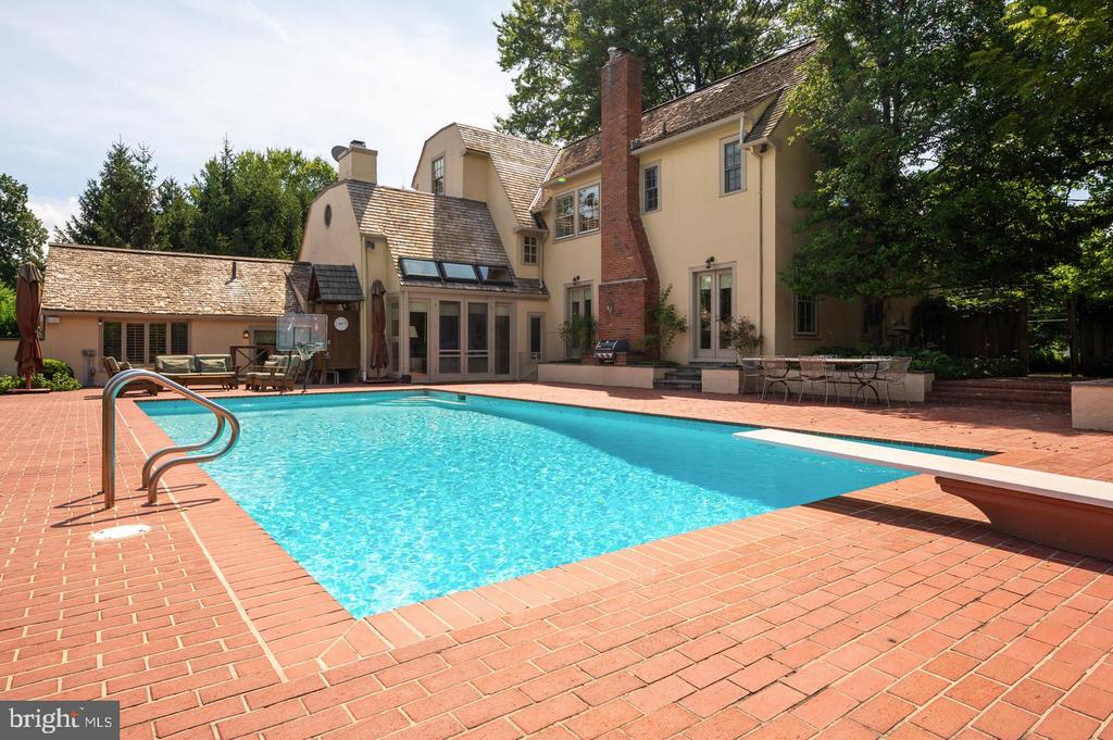 Backyard pool and brick patio beside a large home, with outdoor seating and mature trees.