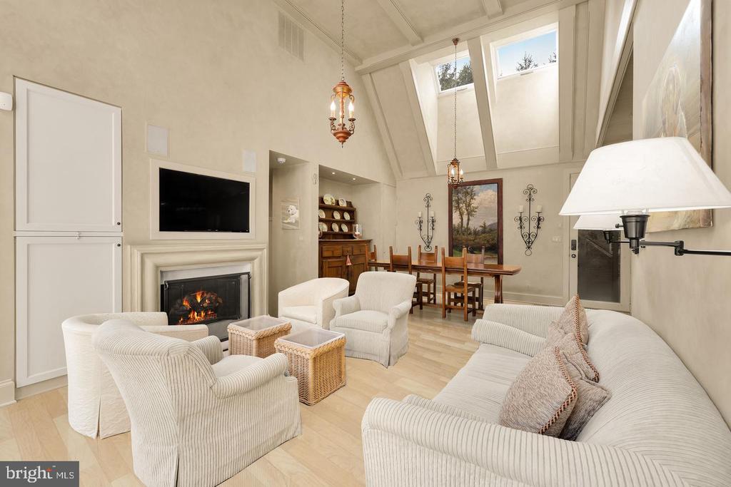 Living room with a fireplace, vaulted skylit ceiling, and pale upholstered seating beside the dining area.