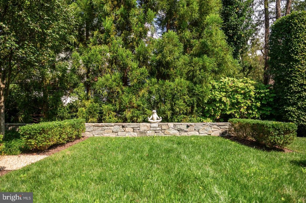 Landscaped garden with a green lawn, low stone wall, and small statue beneath mature trees.