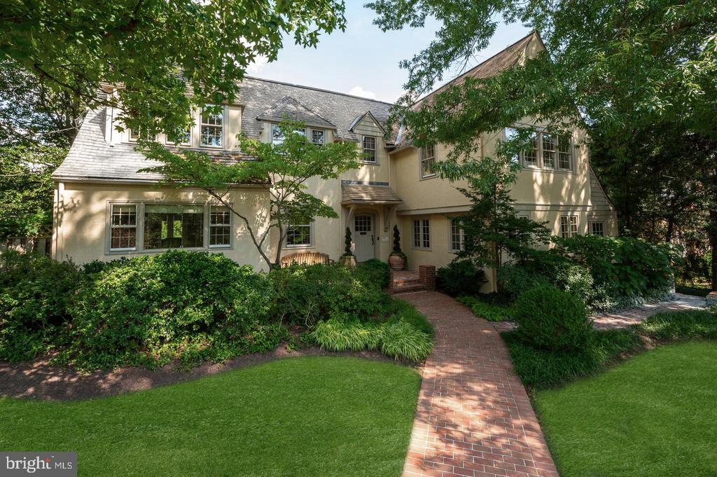 Two-story cream house with a gray shingle roof, framed by mature trees and a brick walkway.