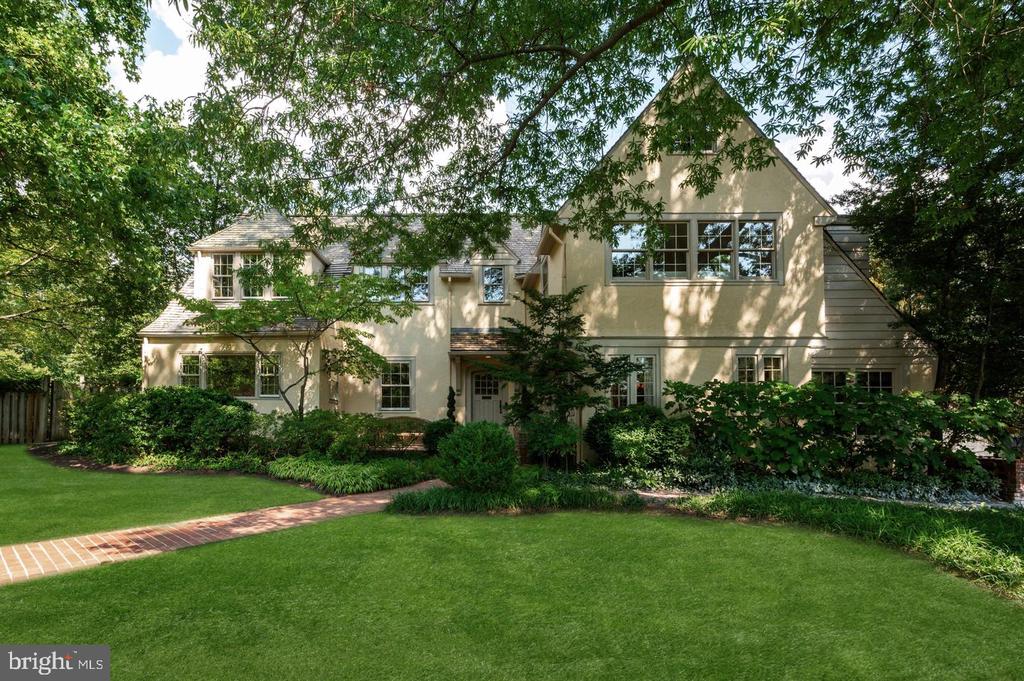 Pale stucco house framed by mature trees, with a broad lawn and brick walkway.