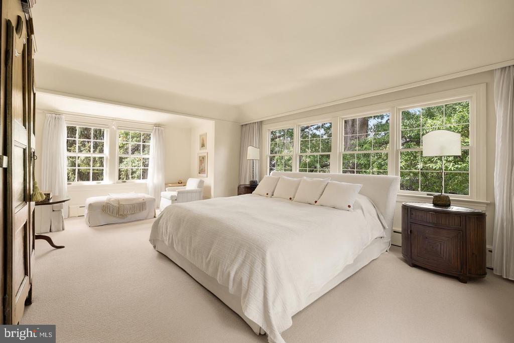 Primary bedroom with a large white bed, sitting area, and wide windows overlooking leafy trees.