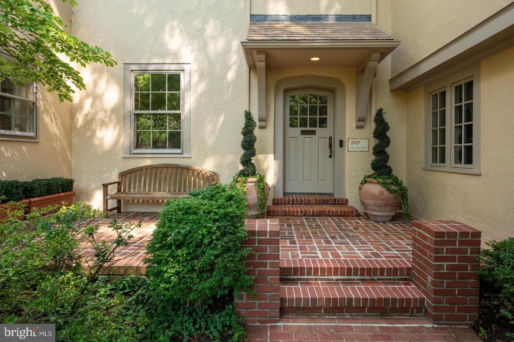 Cream-colored home entrance with red brick steps, a recessed gray door, wooden bench, and landscaped courtyard.