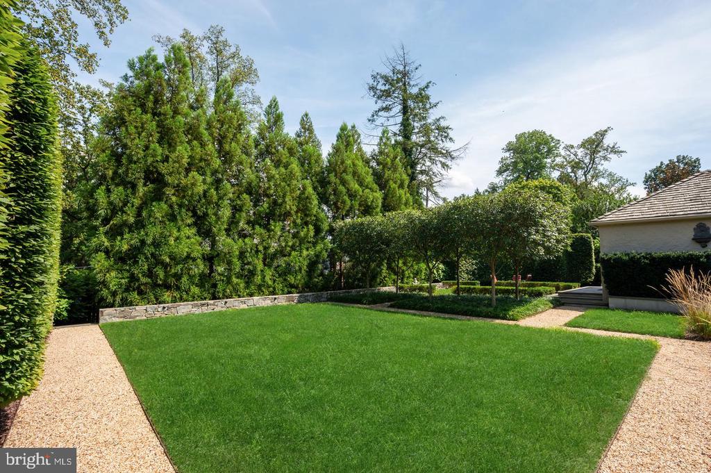 Backyard lawn bordered by gravel paths, mature trees, and tall hedges.