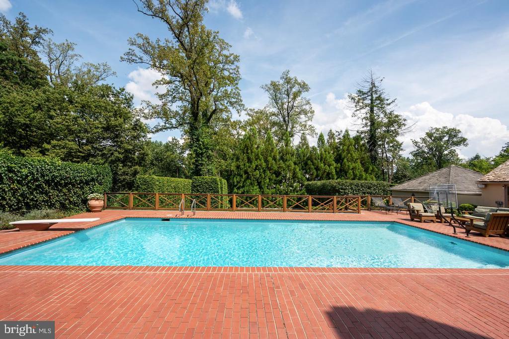 Backyard swimming pool with a broad brick patio, wooden railing, and mature trees.