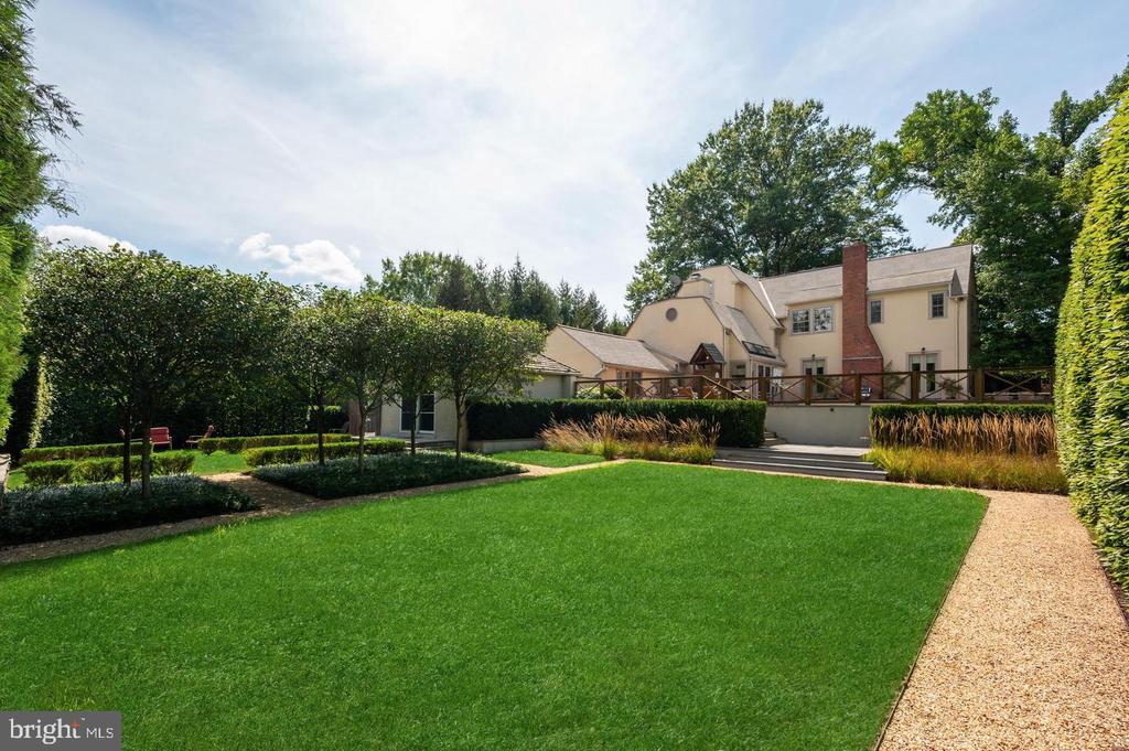 Beige multi-level house behind a broad lawn, landscaped gardens, and a raised wooden deck.