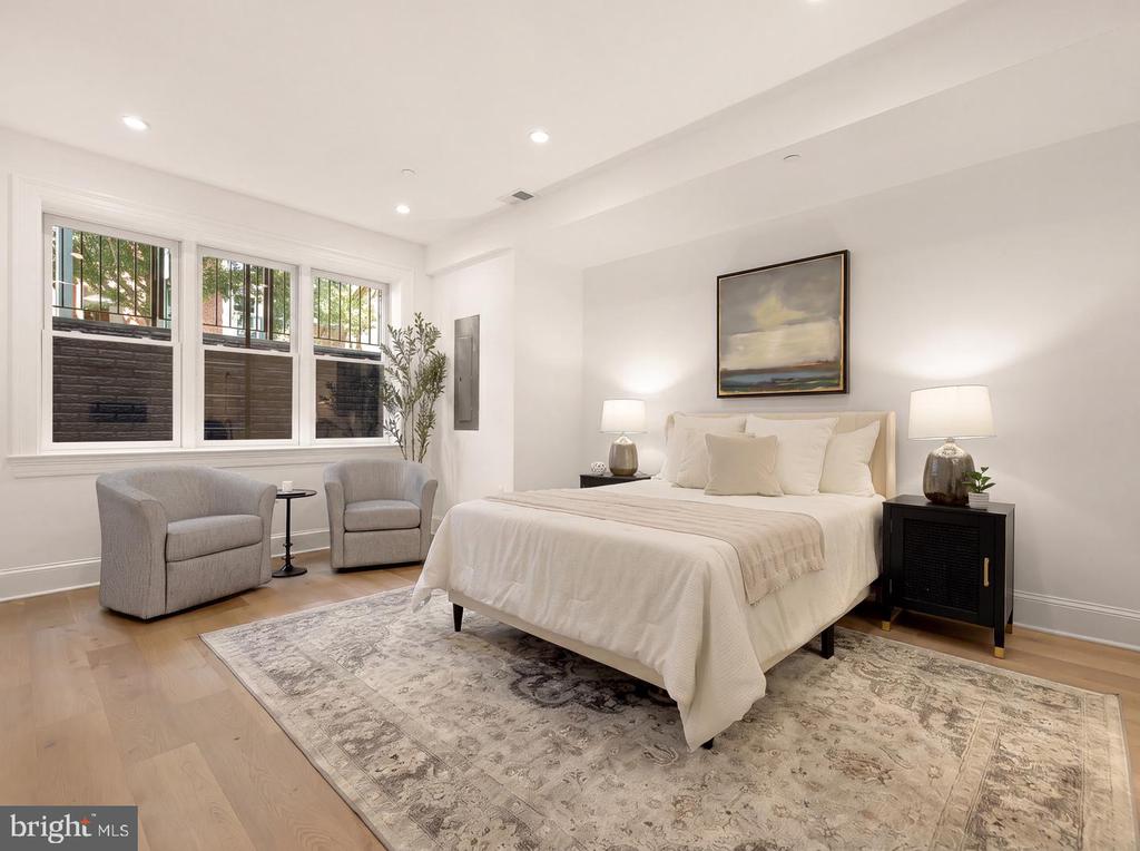 Staged bedroom with a white bed and gray armchairs beside wide windows.