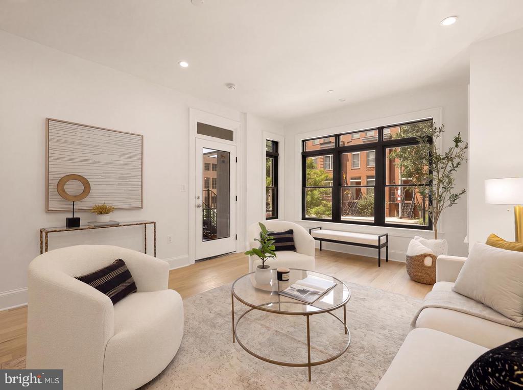 Staged living room with white seating, a glass coffee table, and large windows overlooking brick buildings.