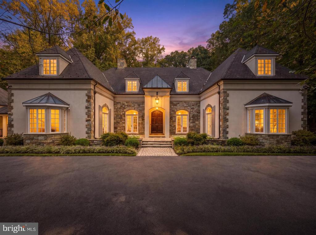 Symmetrical stone-and-stucco house with glowing windows, dormers, and a central arched entrance at dusk.