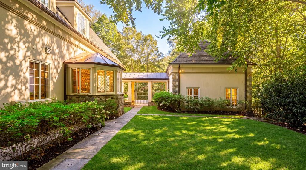 Home’s rear courtyard with a stone walkway, connecting glass passage, and shaded lawn.