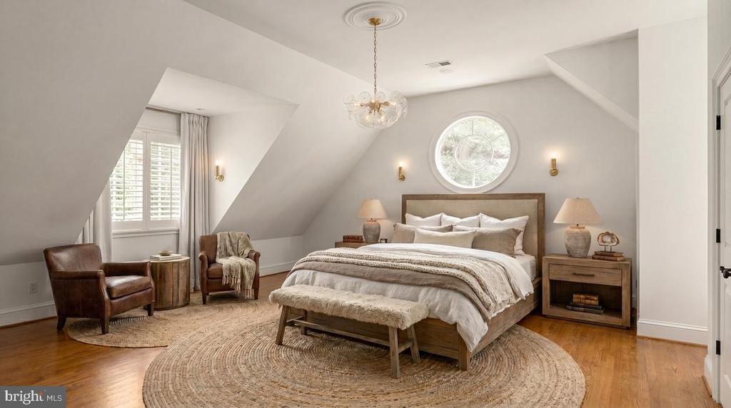 Attic bedroom with a round window above the bed, sloped ceilings, and two leather chairs.