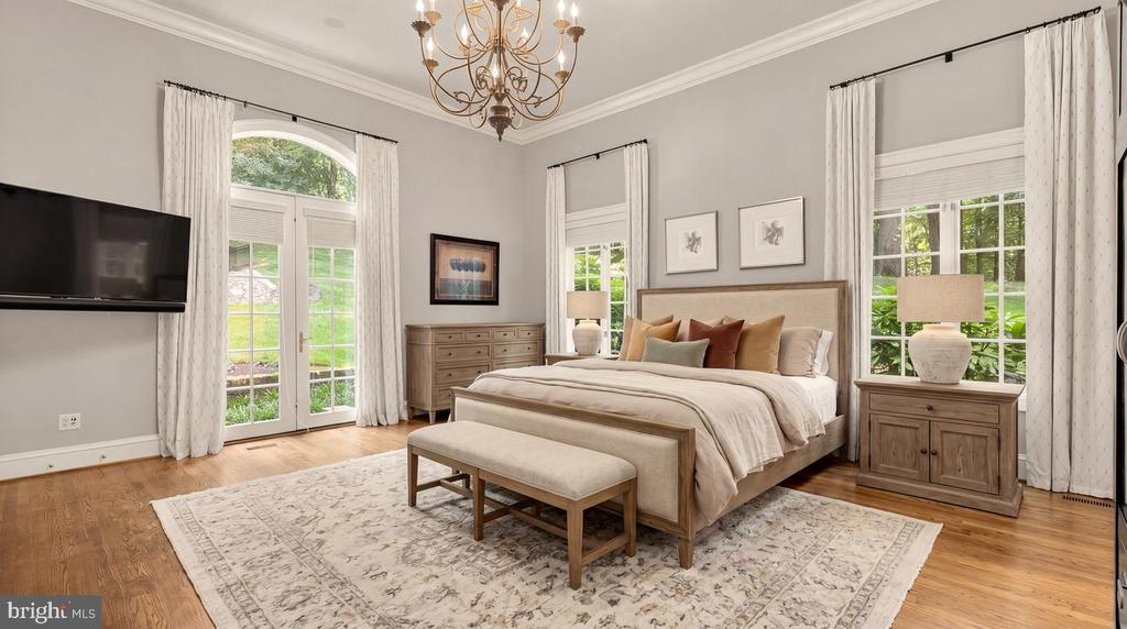 Spacious bedroom with a king bed, upholstered bench, chandelier, and tall windows overlooking greenery.