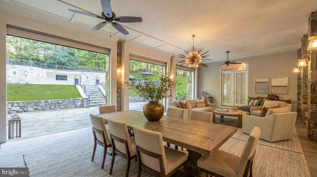 Open-plan dining and living room with a long wood table, stone fireplace, and large windows overlooking a patio.