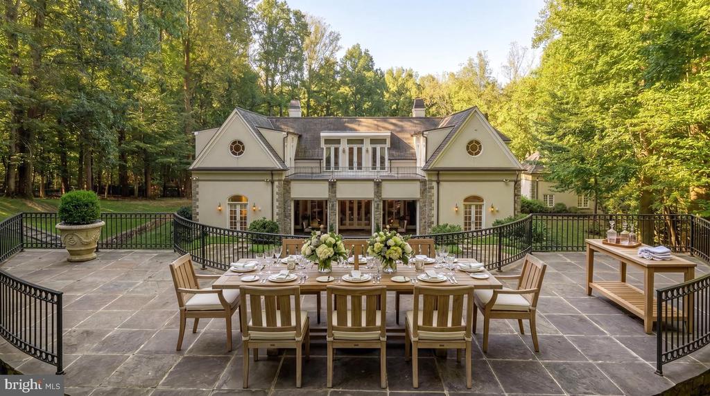 Large cream home beyond a furnished stone terrace, surrounded by mature trees.