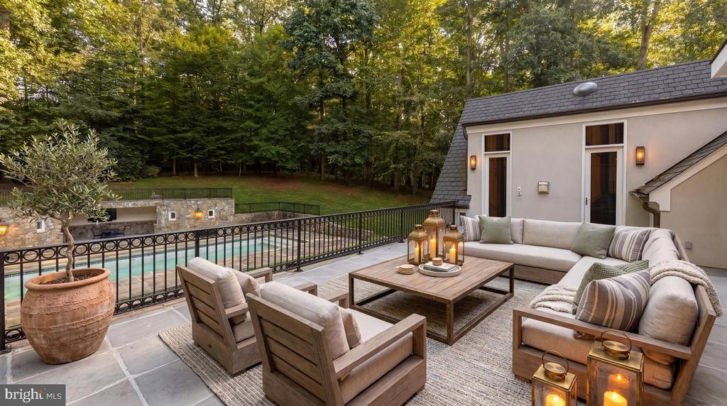 Furnished stone patio with lounge seating and lanterns beside a pool and wooded yard.