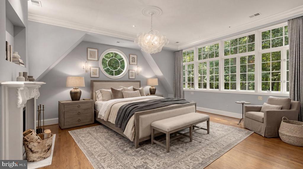 Bedroom with a gray upholstered bed, fireplace, round window, and large windows overlooking trees.