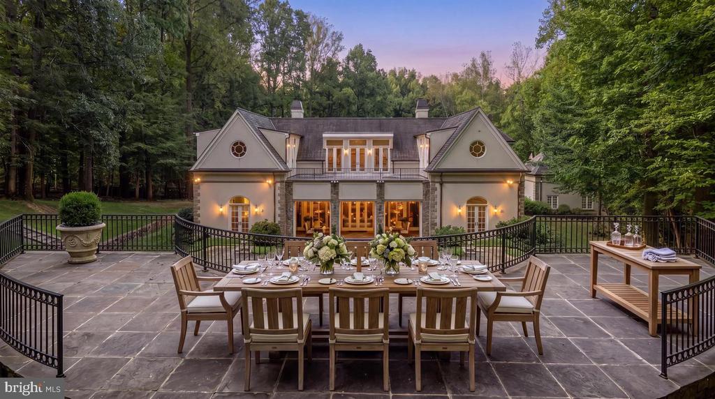 Home’s rear exterior beyond a stone terrace set for outdoor dining, surrounded by trees.