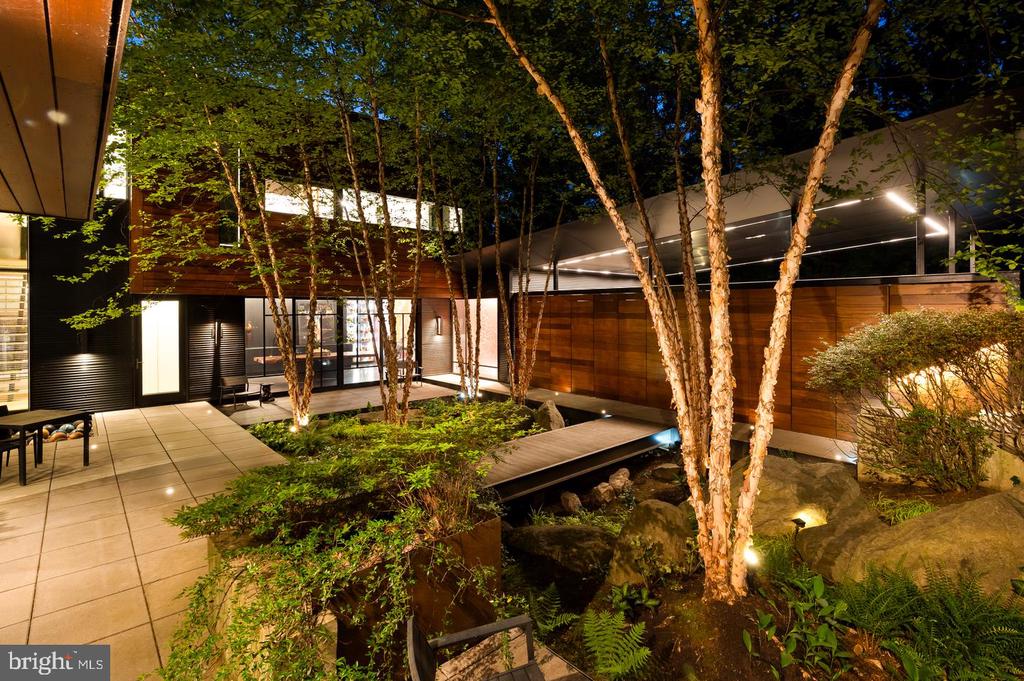 Modern home courtyard with illuminated trees, a tiled walkway, and a rock garden pond.