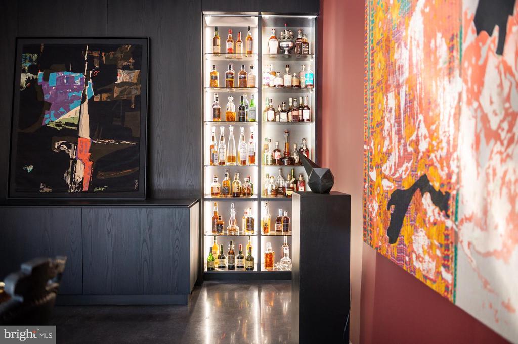 Interior bar with a backlit liquor cabinet, dark cabinetry, and colorful abstract artwork.