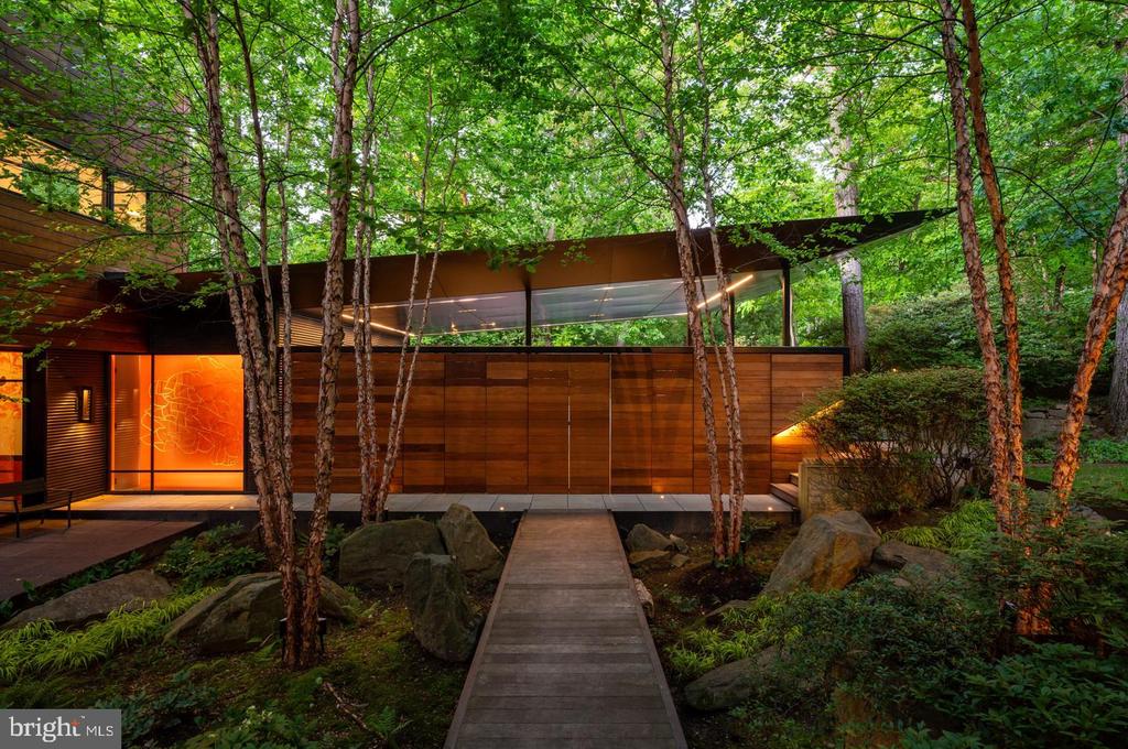 Modern wood-and-glass home among trees, approached by a boardwalk through a rocky garden.