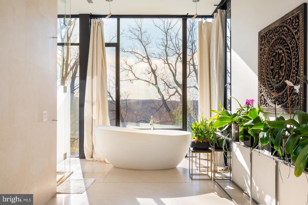 Sunlit bathroom with a freestanding tub beside floor-to-ceiling windows and leafy plants.