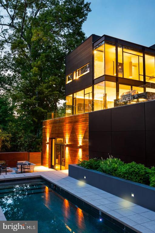 Modern glass-and-panel home glowing at dusk beside a swimming pool and tree-lined patio.