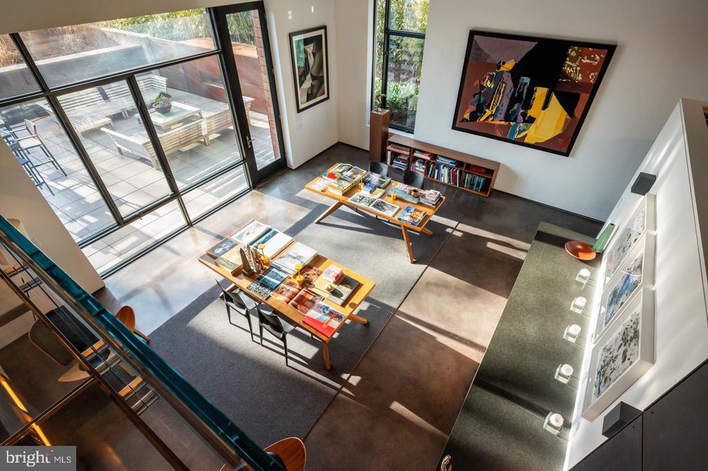 Modern living room with large glass patio doors, colorful artwork, and low tables covered in books and magazines.