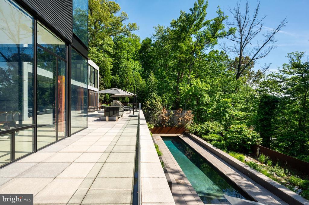 Modern glass home beside a long narrow pool and paved terrace, surrounded by leafy trees.