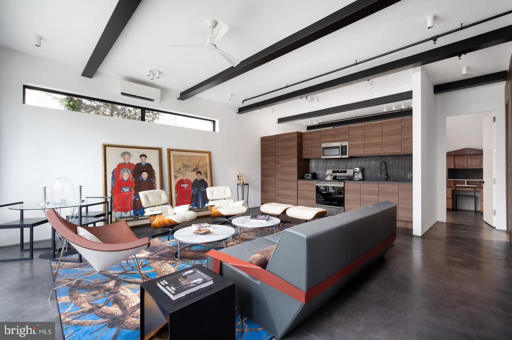Living room with exposed beams, sculptural seating, colorful rug, and an open wood-cabinet kitchen.