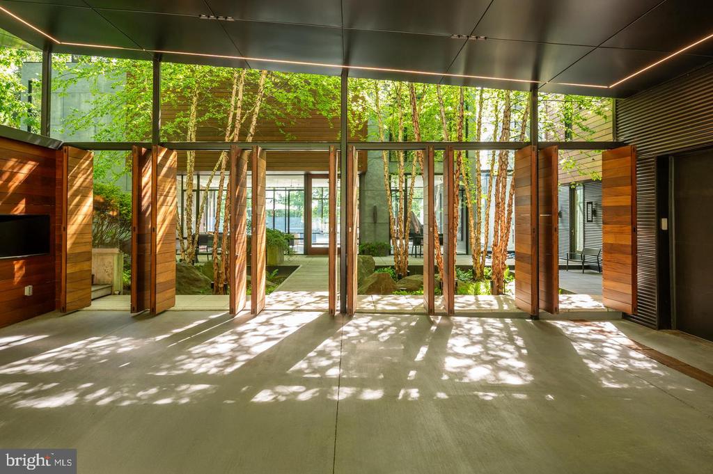 Modern entry hall with open wood-paneled doors, glass walls, and a tree-filled courtyard beyond.