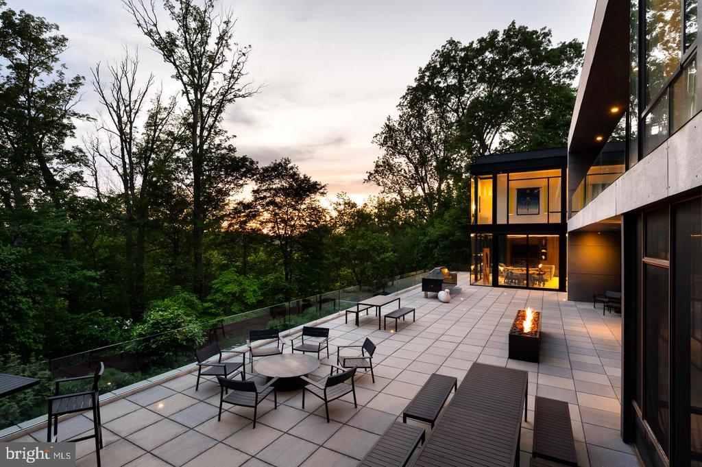 Modern home’s spacious tiled terrace with glass railing, outdoor seating, and a fire feature at dusk.