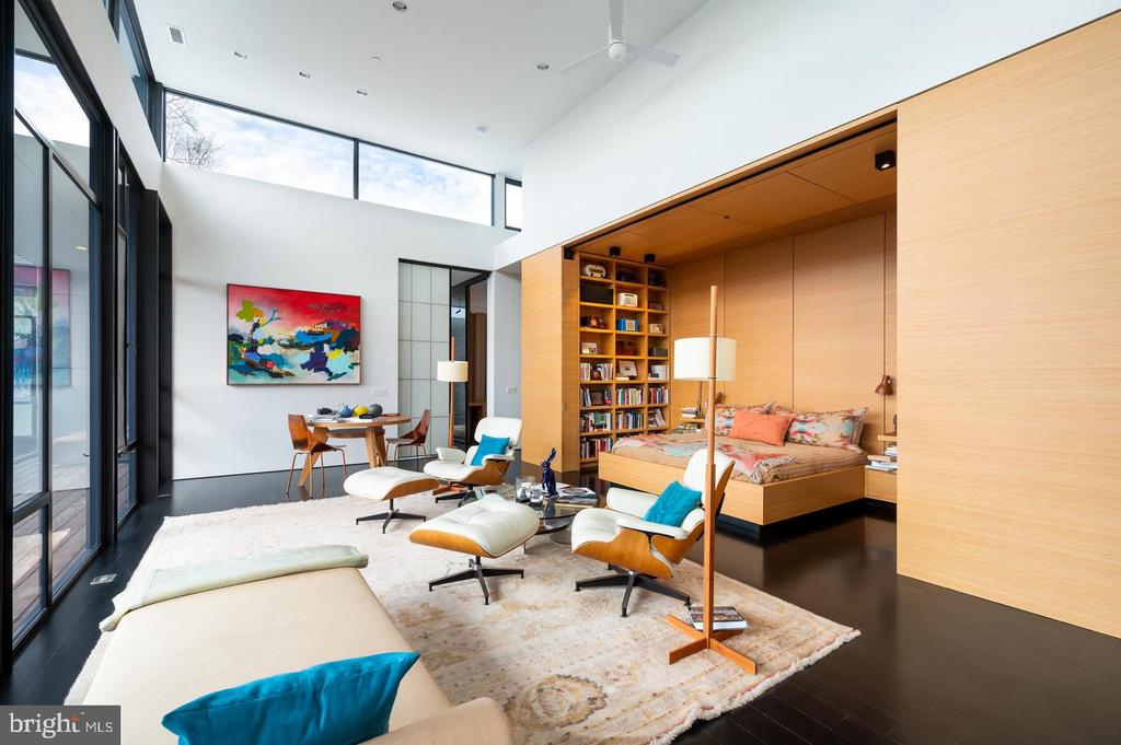 Modern living room with a wood-framed sleeping nook, lounge chairs, and tall windows.