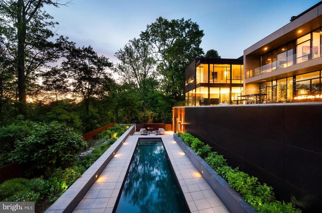 Modern glass home with a narrow lap pool and landscaped grounds at dusk.
