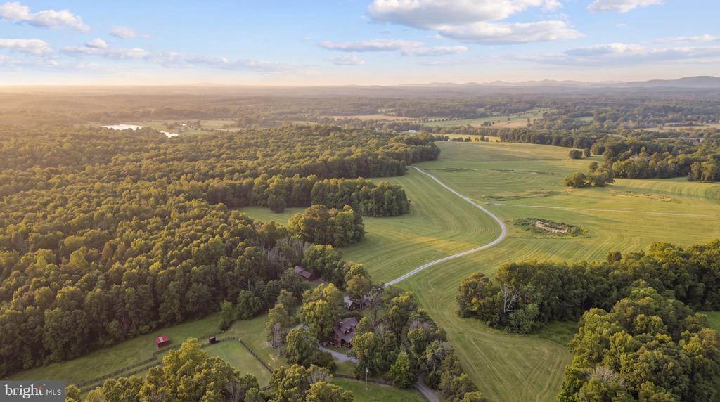 Aerial view of wooded countryside and green fields with a winding driveway and scattered buildings.