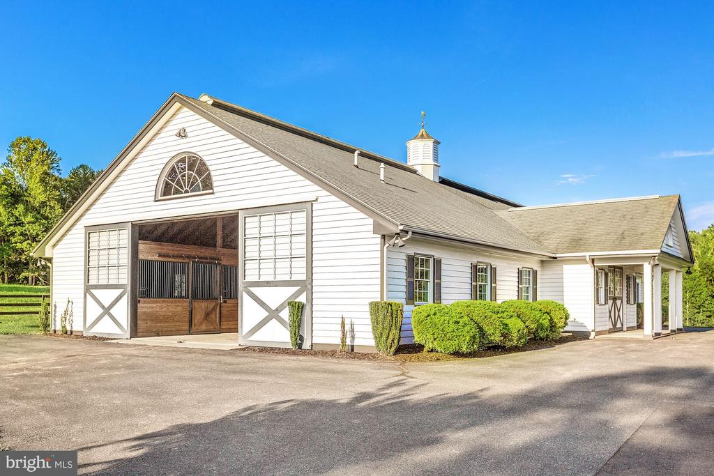 White stable with open horse-stall doors, a cupola, and clipped shrubs beside the paved drive.