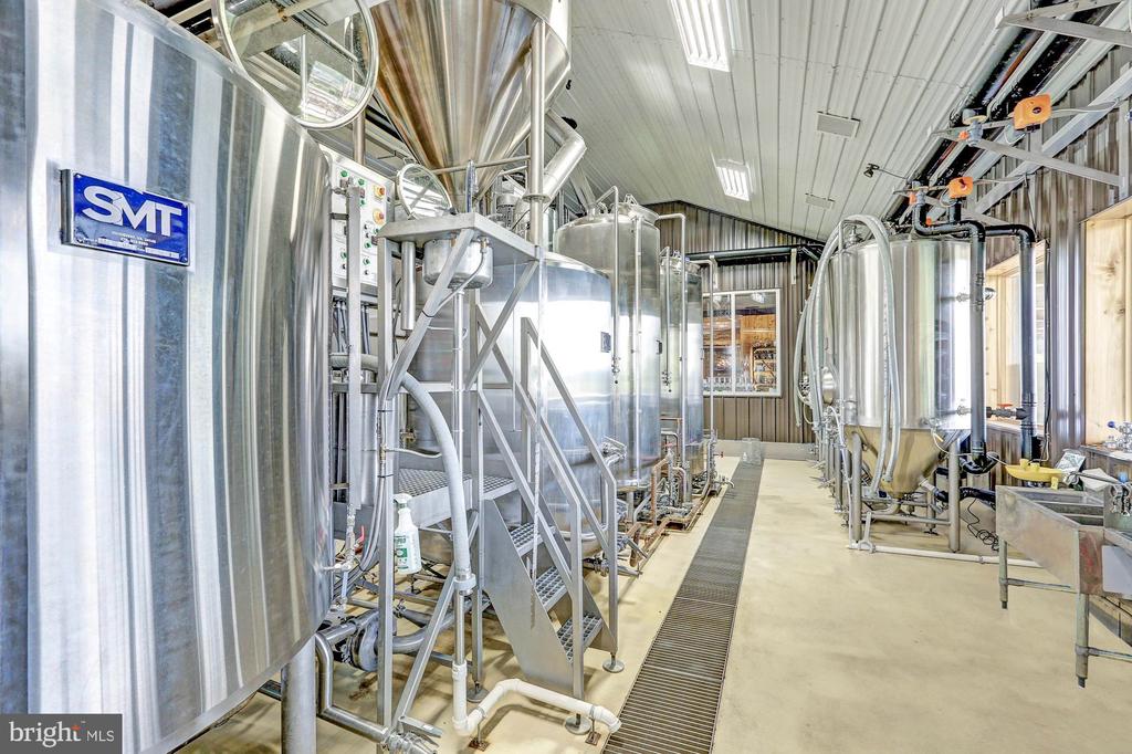 Brewery production room with stainless-steel tanks, overhead pipes, and a central floor drain.