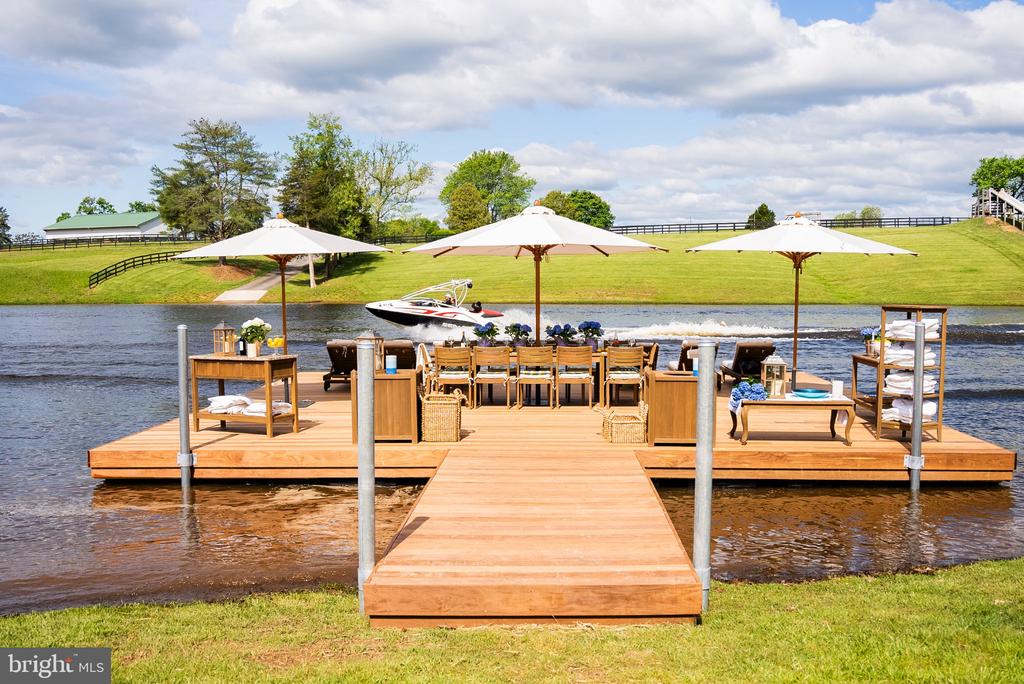 Furnished lakeside dock with a dining table, white umbrellas, and a boat on the water.