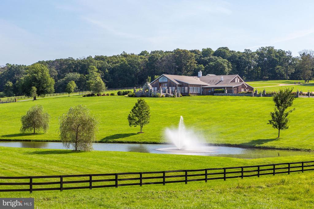Large home beyond a pond with a fountain, manicured lawn, black fence, and wooded backdrop.